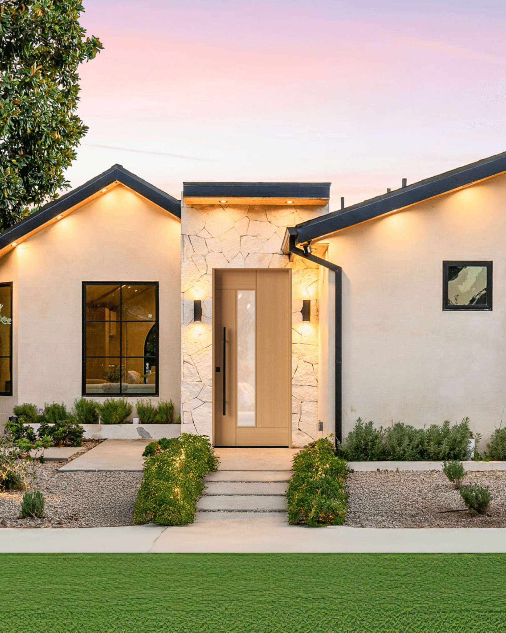 Modern single-story home exterior at dusk with white stucco walls, black trim, and a stone entryway. A light wood DSA modern Sierra front door with a vertical glass panel is centered between two peaked rooflines, with warm exterior lighting illuminating the facade and landscaped gravel yard with low shrubs and a concrete walkway.
